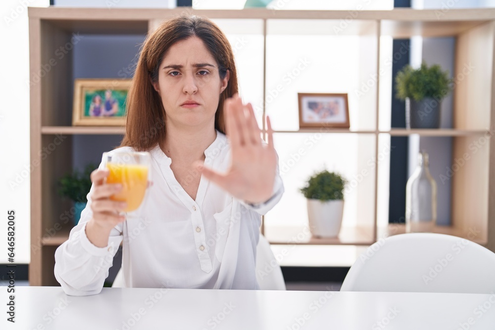 Brunette woman drinking glass of orange juice doing stop sing with palm of the hand. warning expression with negative and serious gesture on the face. Brunette woman drinking glass of orange juice doing stop sing with palm of the hand. warning expression with negative and serious gesture on the face.