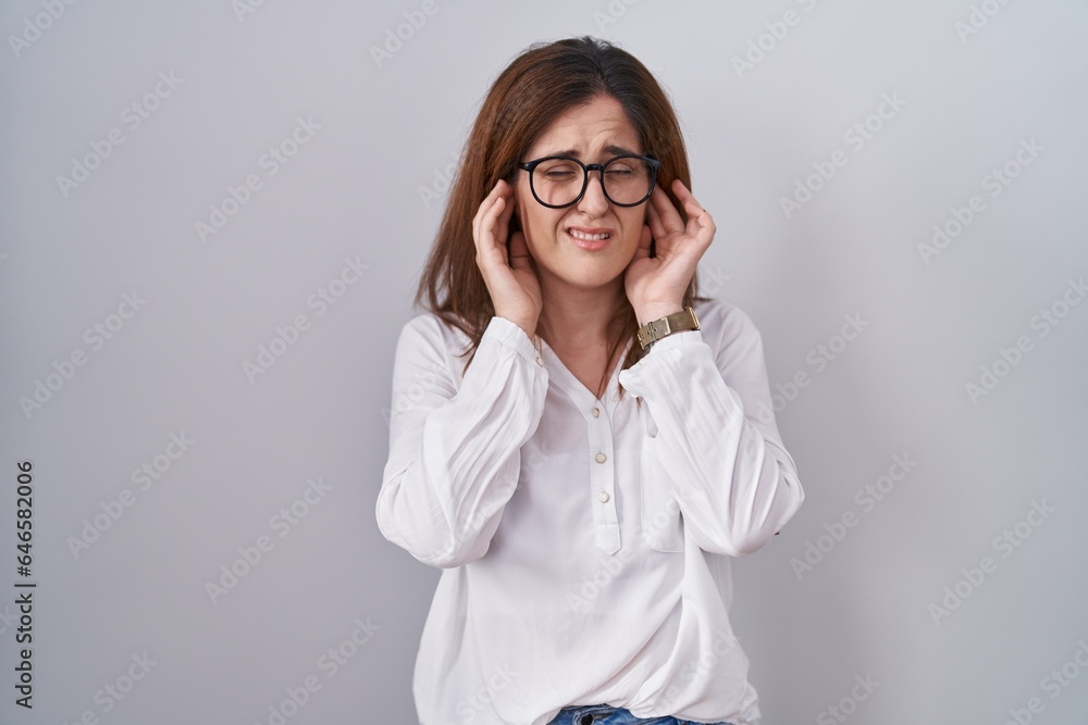 Brunette woman standing over white isolated background covering ears with fingers with annoyed expression for the noise of loud music. deaf concept. Brunette woman standing over white isolated background covering ears with fingers with annoyed expression for the noise of loud music. deaf concept.