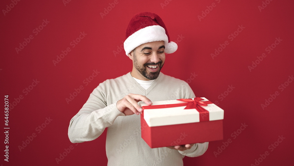 Young hispanic man wearing christmas hat unpacking gift over isolated red background Young hispanic man wearing christmas hat unpacking gift over isolated red background