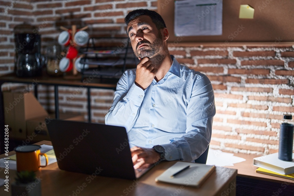 Hispanic man with beard working at the office at night with hand on chin thinking about question, pensive expression. smiling with thoughtful face. doubt concept. Hispanic man with beard working at the office at night with hand on chin thinking about question, pensive expression. smiling with thoughtful face. doubt concept.