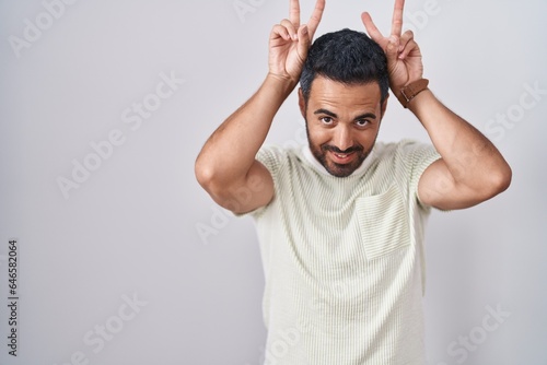 Hispanic man with beard standing over isolated background posing funny and crazy with fingers on head as bunny ears, smiling cheerful