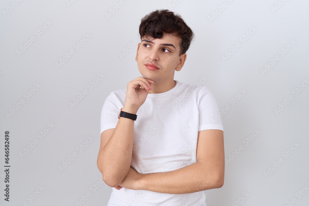 Young non binary man wearing casual white t shirt with hand on chin thinking about question, pensive expression. smiling with thoughtful face. doubt concept. Young non binary man wearing casual white t shirt with hand on chin thinking about question, pensive expression. smiling with thoughtful face. doubt concept.