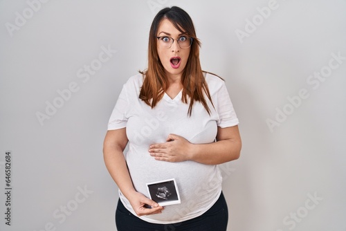 Pregnant woman holding baby ecography in shock face, looking skeptical and sarcastic, surprised with open mouth