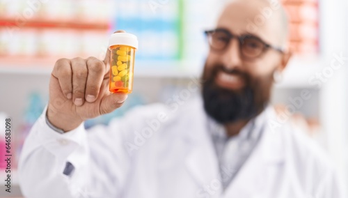 Young bald man pharmacist smiling confident holding pills bottle at pharmacy