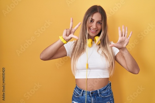 Young blonde woman standing over yellow background wearing headphones showing and pointing up with fingers number eight while smiling confident and happy.