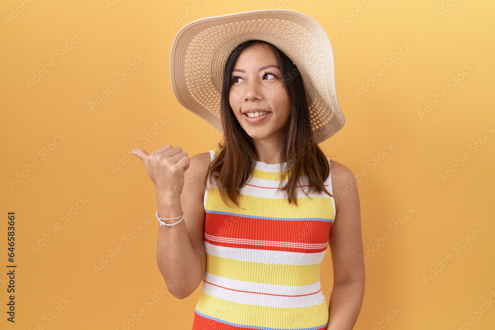 Middle age chinese woman wearing summer hat over yellow background smiling with happy face looking and pointing to the side with thumb up. Middle age chinese woman wearing summer hat over yellow background smiling with happy face looking and pointing to the side with thumb up.