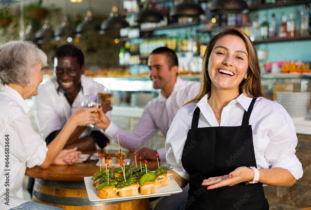 Happy Latin female waiter in uniform pointing hand on the plate with pinchos behind 