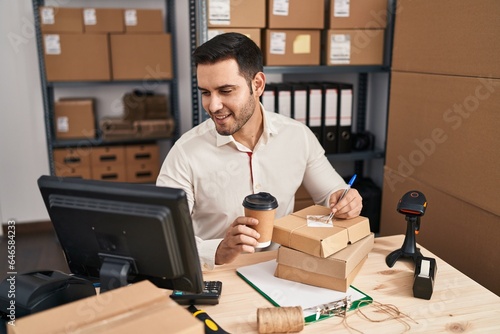 Young hispanic man e-commerce business worker drinking coffee write on package at office