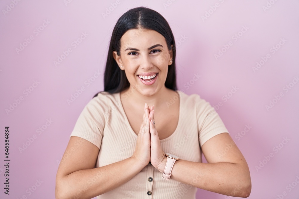 Young hispanic woman standing over pink background praying with hands together asking for forgiveness smiling confident.