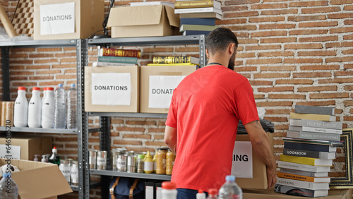 Young hispanic man volunteer standing backwards holding package to donate at charity center