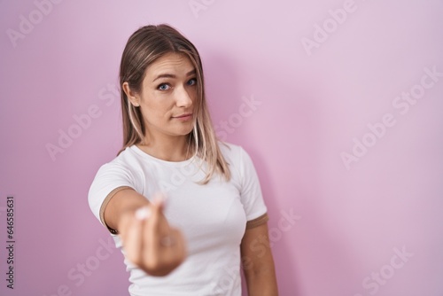 Blonde caucasian woman standing over pink background showing middle finger, impolite and rude fuck off expression