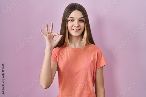 Teenager girl standing over pink background smiling positive doing ok sign with hand and fingers. successful expression.