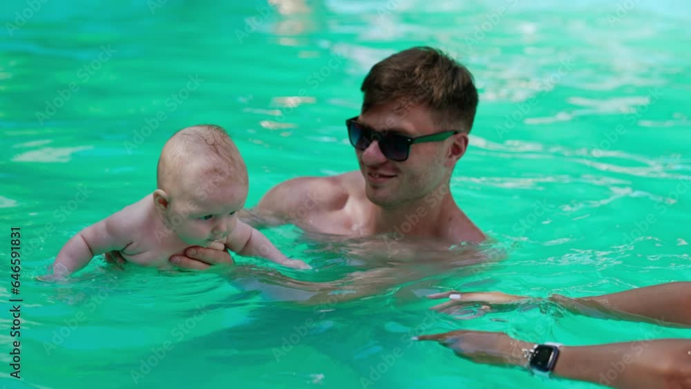 Teaching an infant to swim in the pool. Father is holding a baby and mom stretches hands to him to hold. Teaching an infant to swim in the pool. Father is holding a baby and mom stretches hands to him to hold.