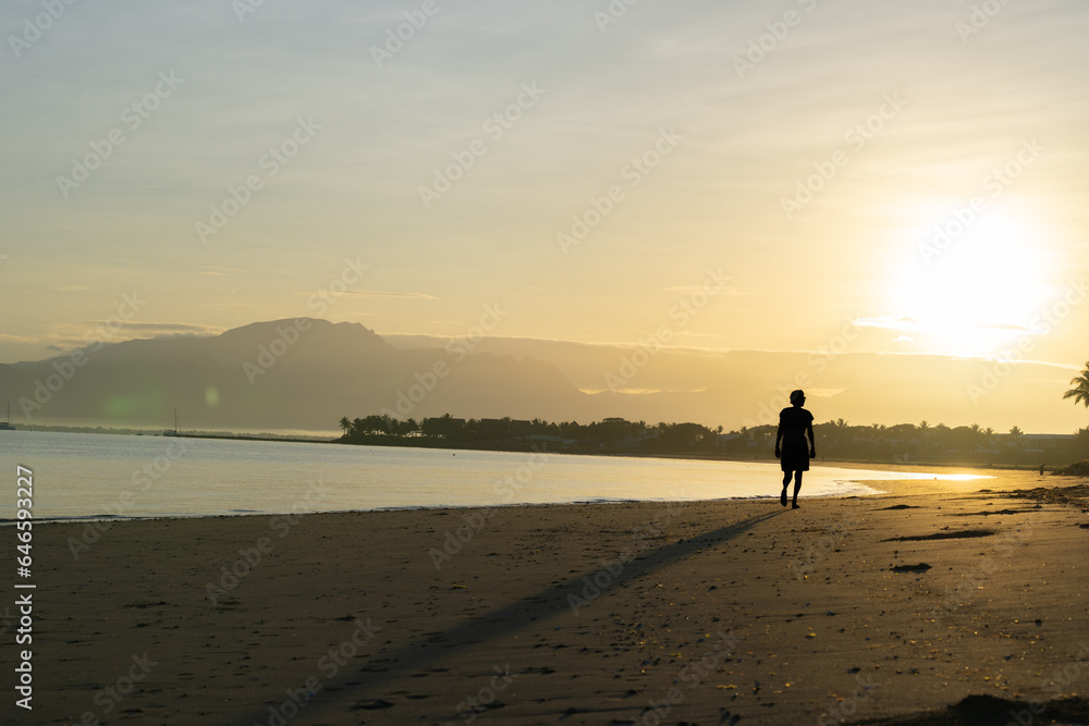 Distant unrecognizable figure of lone person in silhouette walking away on tropical beach at sunrise