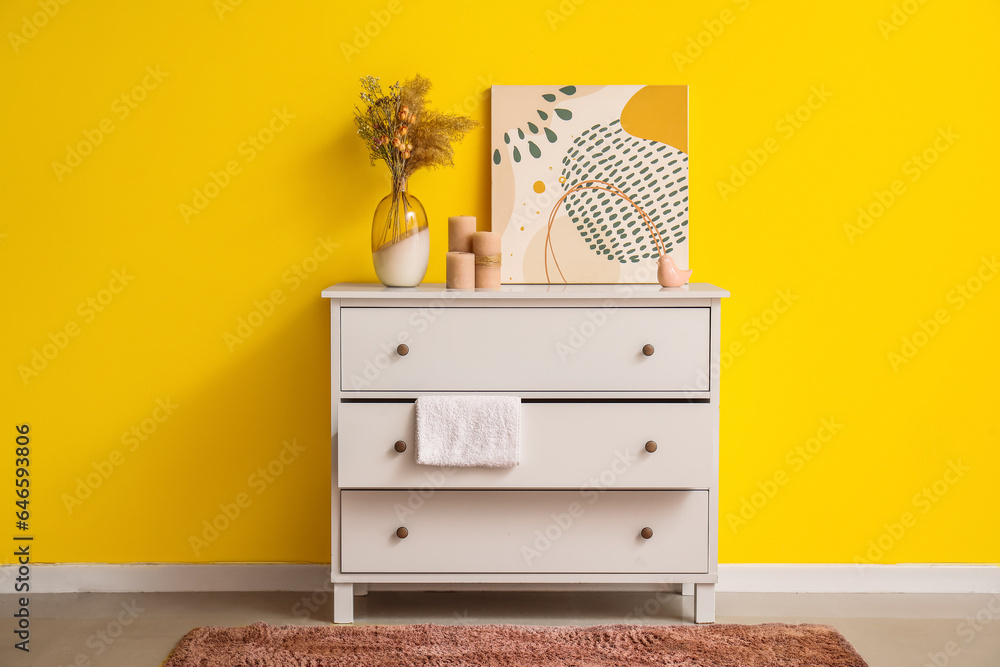 Obraz premium Vase with dried plant and painting on white chest of drawers near yellow wall