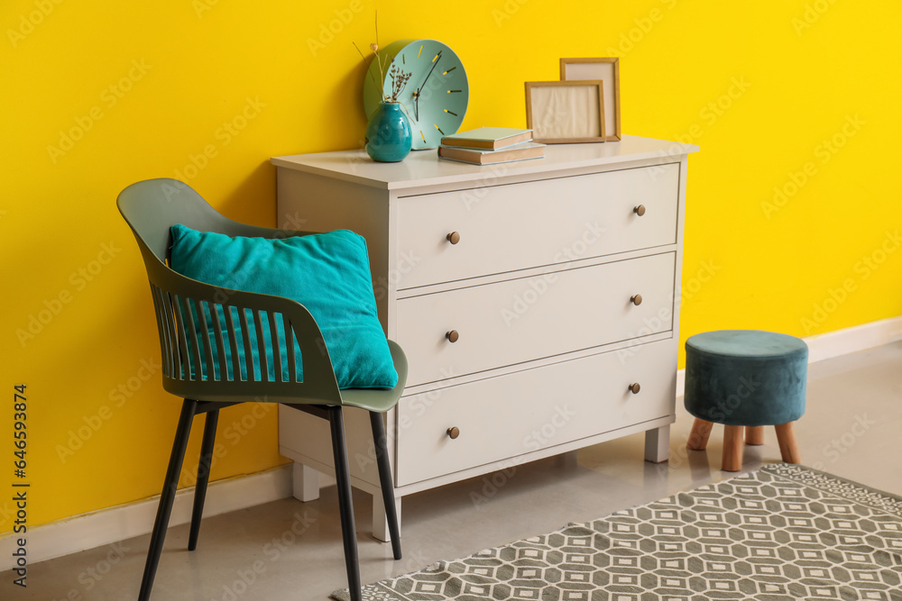 White chest of drawers and stylish chair with cushion near yellow wall White chest of drawers and stylish chair with cushion near yellow wall