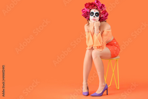 Young woman with painted skull sitting on orange background. Mexico's Day of the Dead (El Dia de Muertos) celebration