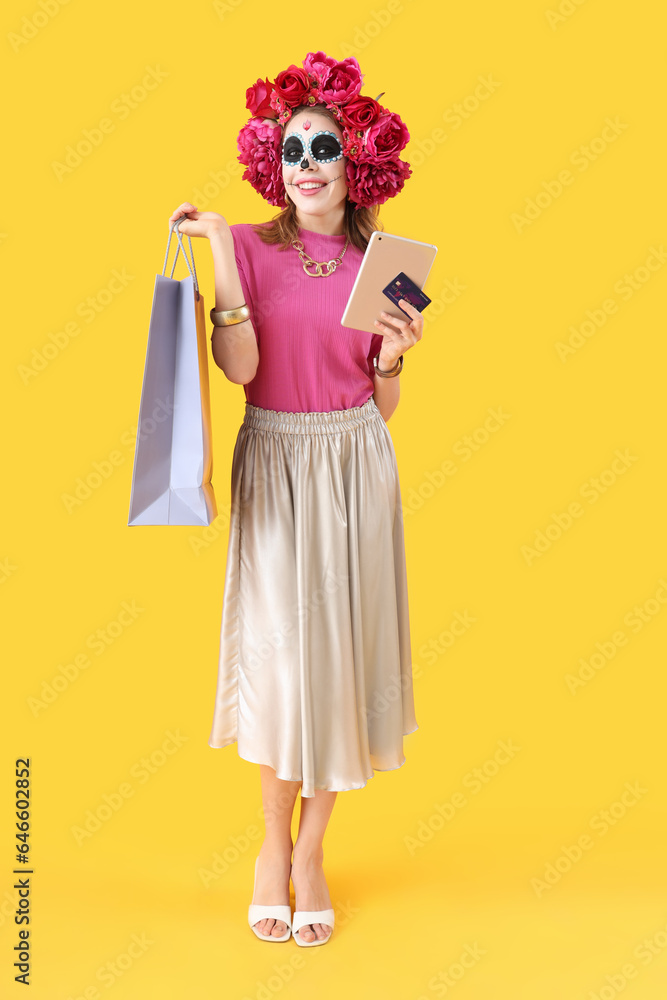 Young woman with tablet computer, credit card and shopping bag on yellow background. Mexico's Day of the Dead (El Dia de Muertos) celebration Young woman with tablet computer, credit card and shopping bag on yellow background. Mexico's Day of the Dead (El Dia de Muertos) celebration