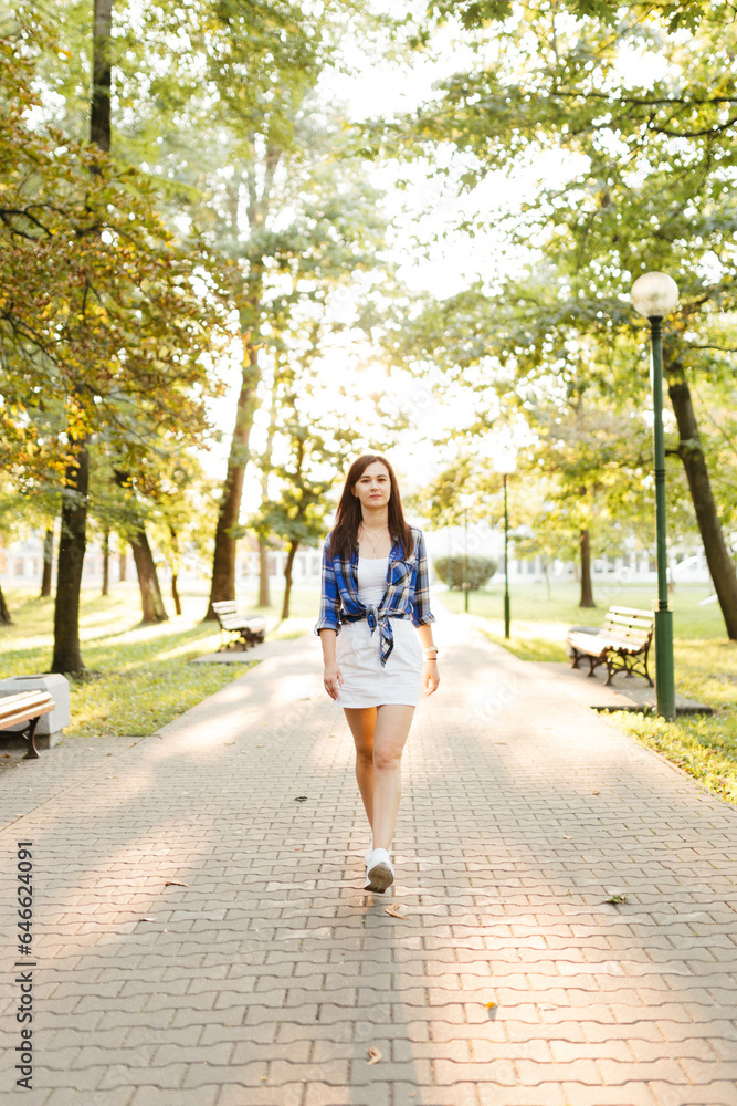 A young beautiful woman in a blue checkered shirt and a white short skirt with black long hair walks and poses in an autumn park. Summer portrait of a beautiful girl on a sunny day A young beautiful woman in a blue checkered shirt and a white short skirt with black long hair walks and poses in an autumn park. Summer portrait of a beautiful girl on a sunny day