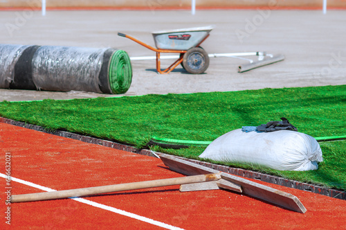 Laying artificial turf on the sports field of the stadium. Green grass carpet rolls and orange soft crumb rubber tracks. Tools and materials on a construction site without people. Sunny day.