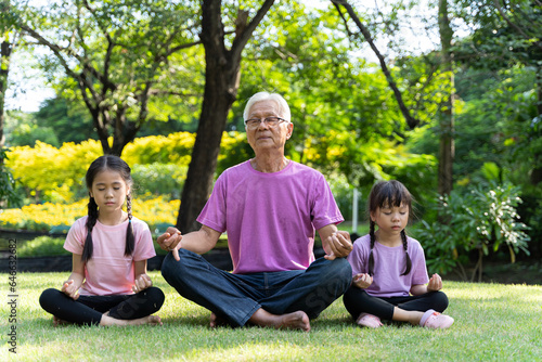 A grandfather and his two daughters sit on the grass in the sunshine doing yoga.