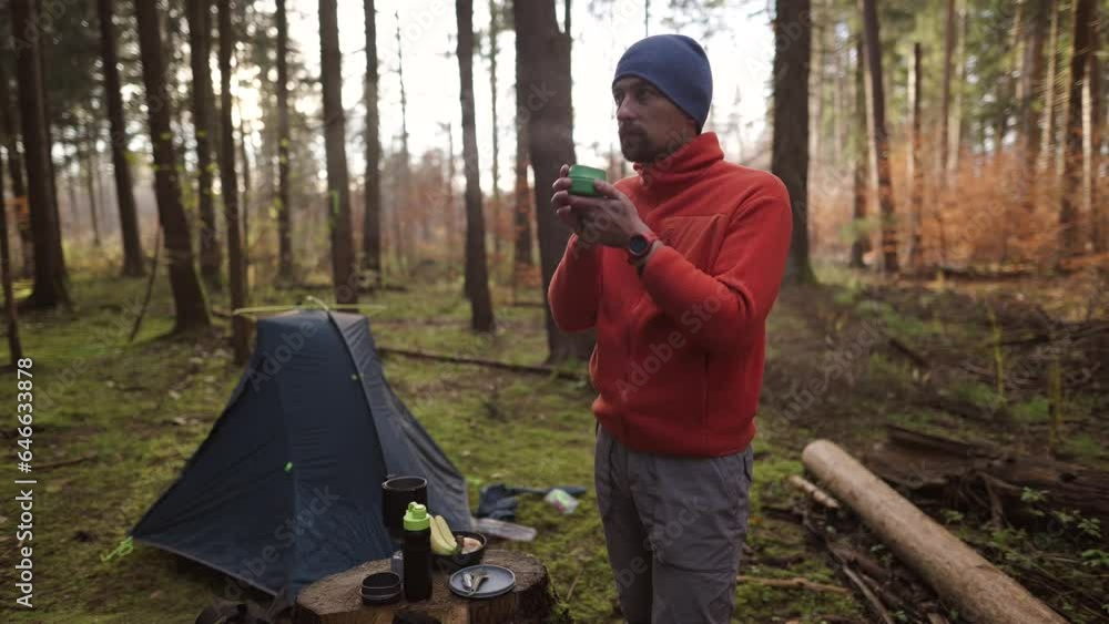 Traveling, adventures, wild nature, exploring. Hiker drinking hot beverage from cup in autumn woods while hiking in front of camping tent. Heating with tea during a backpacking trip in cold weather. Traveling, adventures, wild nature, exploring. Hiker drinking hot beverage from cup in autumn woods while hiking in front of camping tent. Heating with tea during a backpacking trip in cold weather.
