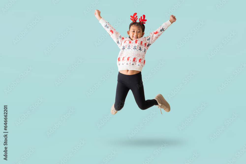 Cheerful young Asian girl wearing a Christmas sweater with reindeer horns, Happy smiling dance jumping have fun full body portrait, isolated on pastel plain light blue background.