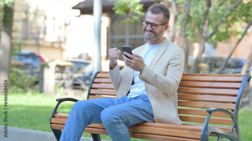 Obraz premium Middle Aged Man Celebrating Success on Smartphone while Sitting Outdoor on a Bench
