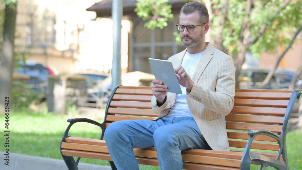 Obraz premium Middle Aged Man using Tablet while Sitting Outdoor on a Bench