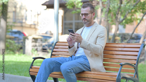 Middle Aged Man using Smartphone while Sitting Outdoor on a Bench