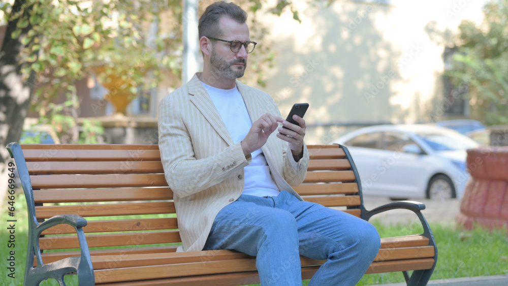 Fototapeta premium Modern Casual Man using Smartphone while Sitting Outdoor on a Bench