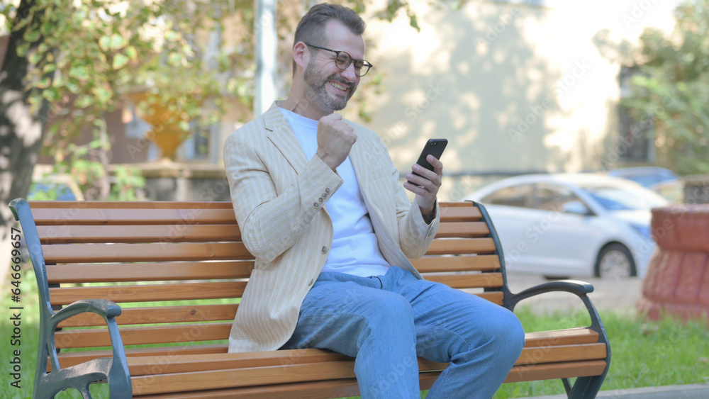 Fototapeta premium Modern Casual Man Celebrating Success on Smartphone while Sitting Outdoor on a Bench