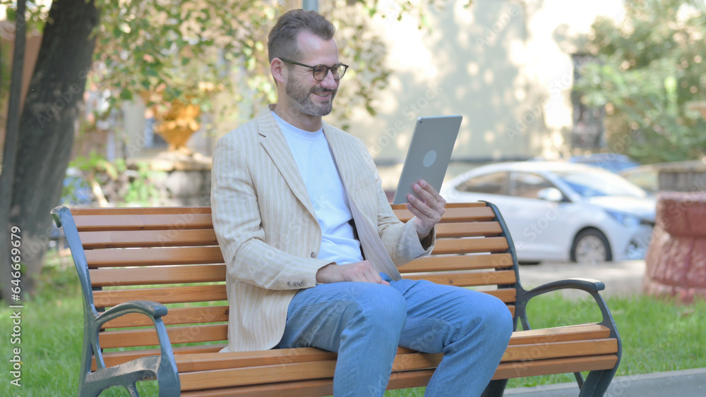 Obraz premium Modern Casual Man Doing Video Chat on Tablet while Sitting Outdoor on a Bench
