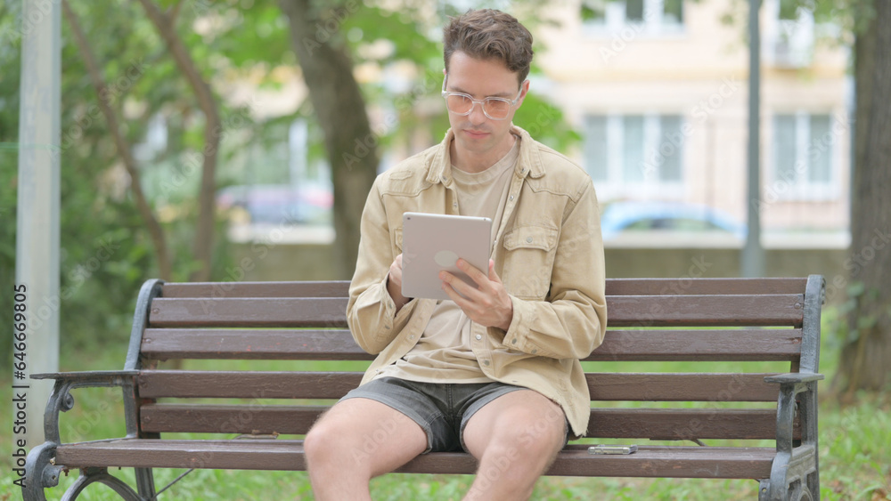 Fototapeta premium Casual Young Man using Tablet while Sitting Outdoor on a Bench
