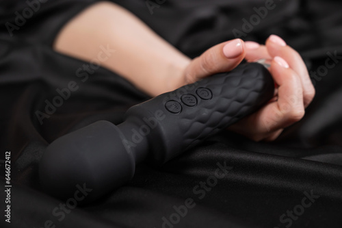 A woman is holding a black vibrator while lying on a black silk sheet.