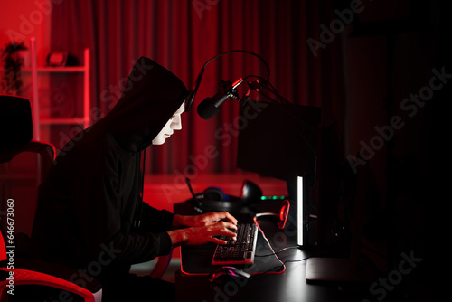 Hoodie player hack game typing and coding with network data online in computer while wearing white mask hide face for cyber attack at security server red room