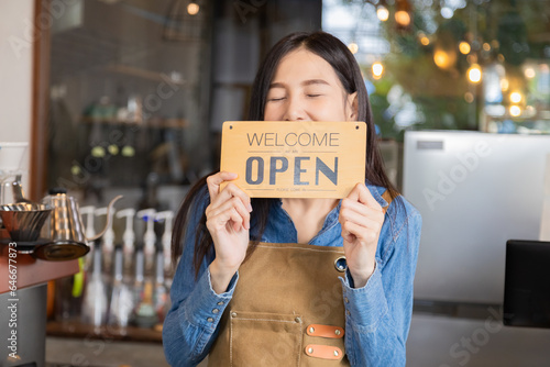 Happy Asian woman coffee business owner holds welcome open signboard ready for opening, young barista employee female standing in front of cafe counter wearing apron successful small business start-up