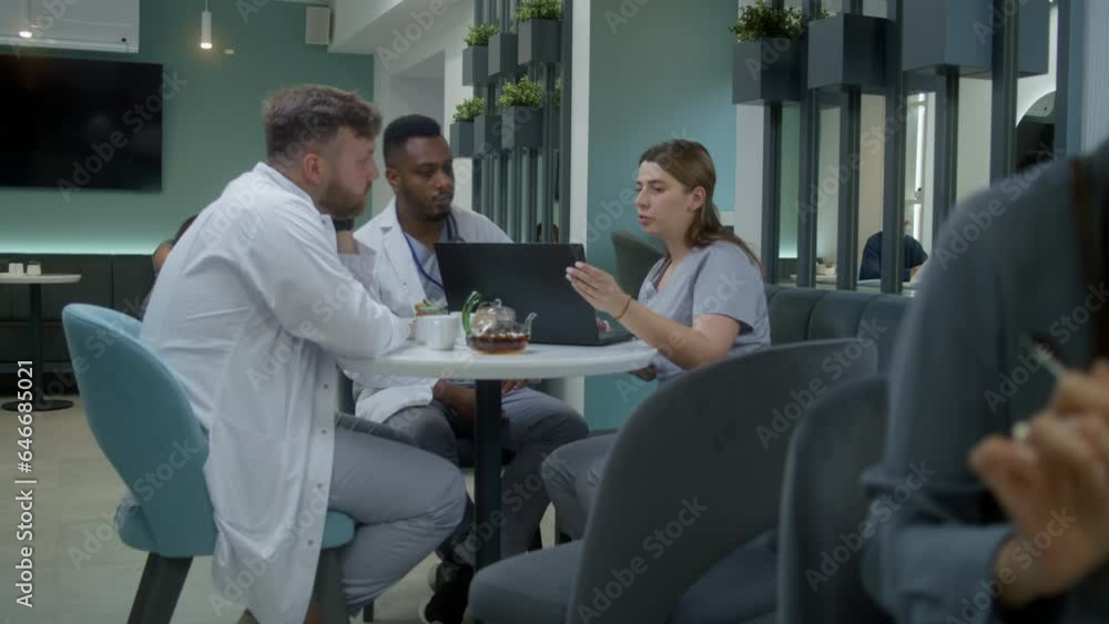 Multiethnic doctors sit in clinic cafe, use laptop. Professional medics talk, examine medical tests results of patients, discuss diagnosis. Medical staff work in hospital or medical center cafeteria.