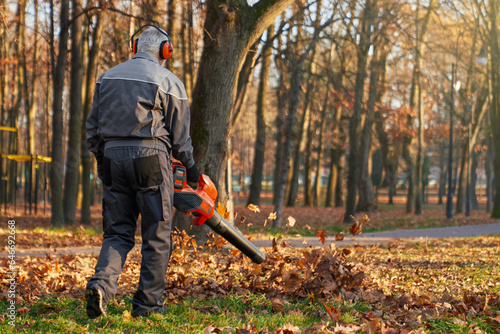 Anonymous male municipal worker cleaning up resting area in autumn day. Back view of man wearing overalls and earmuffs using leaf blower equipment, working outdoor. Concept of seasonal work. photo