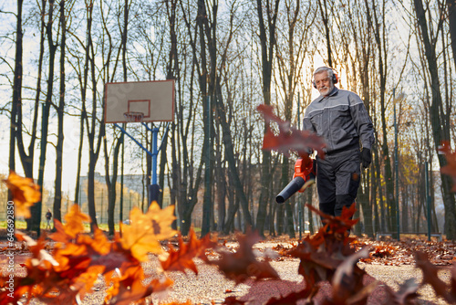 Smiling man in age working with handheld leaf blower, while leaves curling and glowing in pleasant sunlight. Low angle view of worker wearing uniform, blowing out golden leaves in park. Fall concept. photo