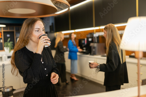 Young women share small talk and drink hot beverages in office canteen. Female employees communicate and sip warm drinks during work break