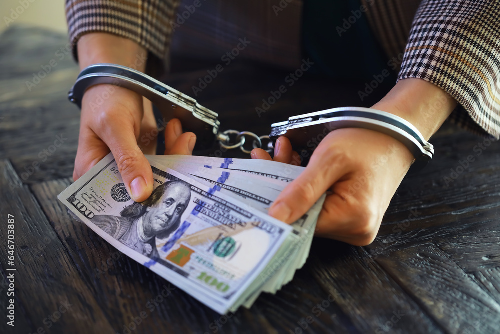 women's handcuffed hands hold dollars on a dark wooden background - obrazy, fototapety, plakaty 