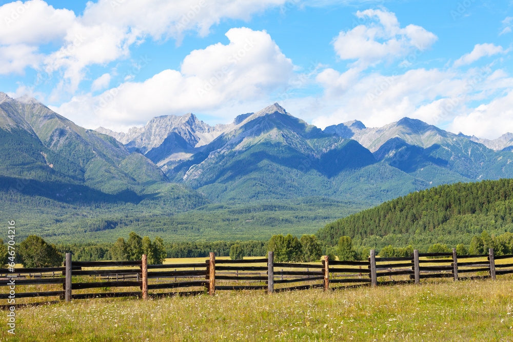 Beautiful mountain landscape with hay meadow behind wooden fence and mountain ridge against background of blue sky with clouds on sunny day. Sayan Mountains, Tunka foothill valley, Buryatia, Siberia