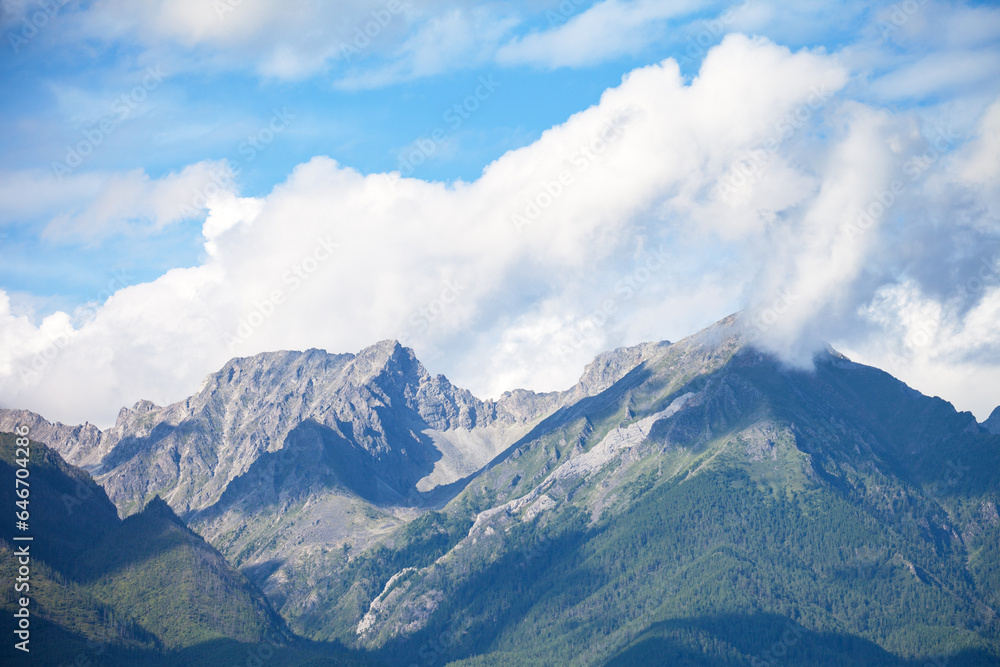 View of mountain range with rocky peaks and low cumulus clouds on sunny summer day. Scenic mountain landscape. Natural background. Eastern Sayan Mountains, Baikal region, Tunka, Buryatia, Siberia