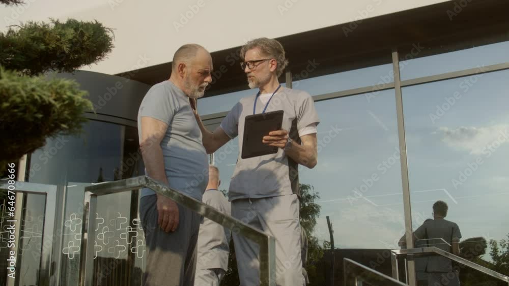 Mature physician with digital tablet in hands stands with aged patient at clinic entrance and discuss his treatment. Health care specialist consults client or makes diagnosis. Medic works outside. Mature physician with digital tablet in hands stands with aged patient at clinic entrance and discuss his treatment. Health care specialist consults client or makes diagnosis. Medic works outside.