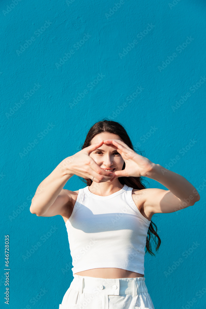 very beautiful young adult woman brunette with long hair in white tight t-shirt with hands making shapes and enjoying smiling angainst blue wall background outdoors isolated very beautiful young adult woman brunette with long hair in white tight t-shirt with hands making shapes and enjoying smiling angainst blue wall background outdoors isolated