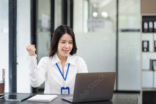 Smiling young asian woman succeeding in business using laptop and computer while doing some paperwork at office.