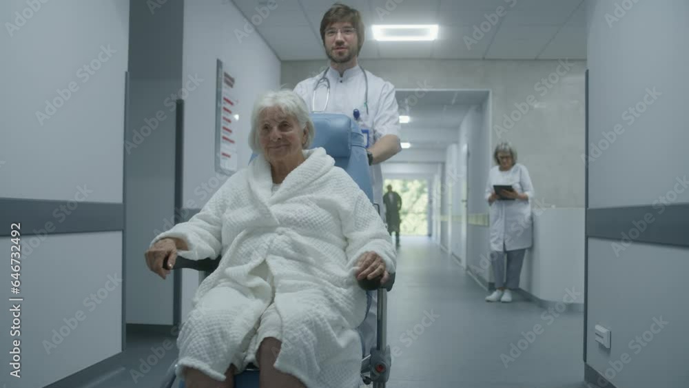 Young doctor in glasses carries senior patient in wheelchair down hospital corridor. Old woman talks to medic and smiles. Mature doctor asks female patient about her health. Medical staff at work. Young doctor in glasses carries senior patient in wheelchair down hospital corridor. Old woman talks to medic and smiles. Mature doctor asks female patient about her health. Medical staff at work.
