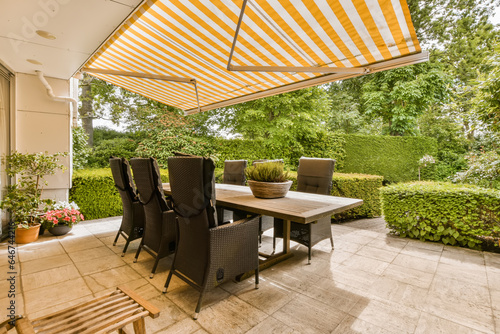 an outdoor dining area with table and chairs under a large yellow striped awning over the patio, surrounded by lush green trees photo