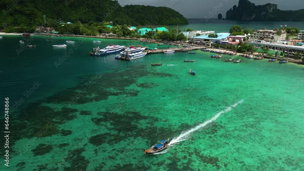 Stunning Aerial Tracking Shot Of A Long Tail Boat Water Taxi Departing The Busy Seaport Village Of Koh Phi Phi, One Of Thailand's Most Visited Islands. Stunning Aerial Tracking Shot Of A Long Tail Boat Water Taxi Departing The Busy Seaport Village Of Koh Phi Phi, One Of Thailand's Most Visited Islands.